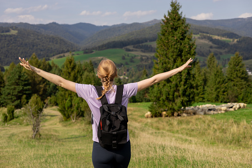 Woman hiker with backpack open arms at mountain peak. Travel and active lifestyle concept. 30s girl enjoying valley view from top of a mountain, outdoor activities, rear view