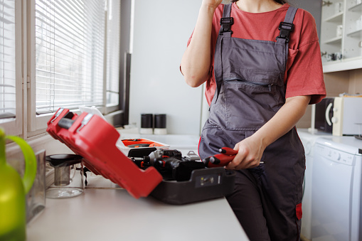 Cropped shot of a handywoman in uniform standing by the opened toolbox, stacking her equipment and attending to clients on the phone