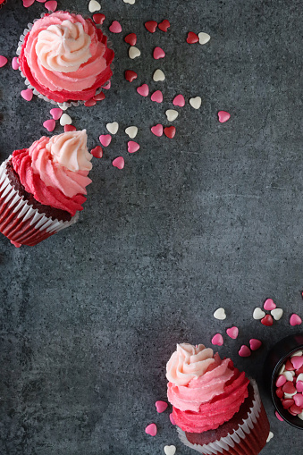 Stock photo showing close-up, elevated view of three, freshly baked, homemade, red velvet cupcakes in paper cake cases with bowl of . The cup cakes have been decorated with swirls of ombre effect pink piped icing and sugar hearts.  Valentine's Day and romance concept.