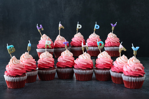 Stock photo showing close-up view of melted, brightly coloured individual, letter candles spelling 'Happy Birthday' stuck into rows of freshly baked, homemade, red velvet cupcakes in paper cake cases. The cup cakes have been decorated with swirls of ombre effect pink piped icing.