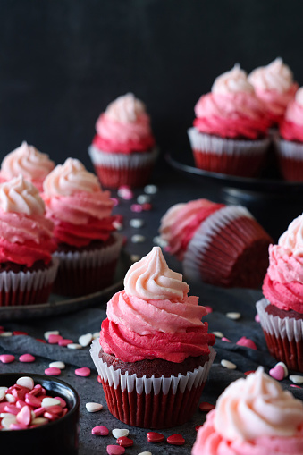 Stock photo showing close-up view of a batch of freshly baked, homemade, red velvet cupcakes in paper cake cases on and surrounding a plate and cake stand. The cup cakes have been decorated with swirls of ombre effect pink piped icing and sugar hearts.  Valentine's Day and romance concept.