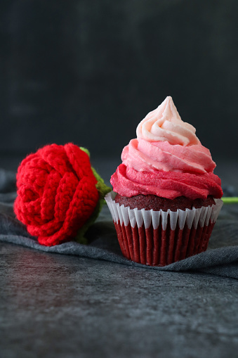 Stock photo showing close-up view of a single freshly baked, homemade, red velvet cupcake in paper cake case with crochet rose with red petals, green stem and leaves on a grey muslin cloth. The cup cake has been decorated with a swirl of ombre effect pink piped icing. Valentine's Day and romance concept.
