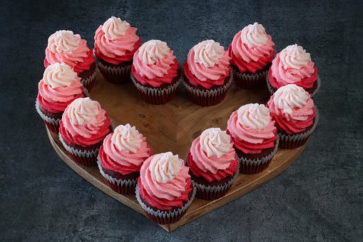 Stock photo showing close-up, elevated view of a batch of freshly baked, homemade, red velvet cupcakes in paper cake cases on and surrounding a heart-shaped, wooden chopping board. The cup cakes have been decorated with swirls of ombre effect pink piped icing and sugar hearts.  Valentine's Day and romance concept.