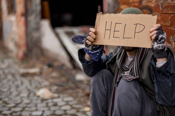 Young Homeless Man Hides His Face With Help Sign While Sitting on a Street . Homelessness Social Issues Concept.