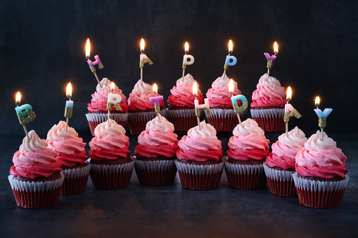 Stock photo showing close-up view of melting, illuminated, brightly coloured individual, letter candles spelling 'Happy Birthday' stuck into rows of freshly baked, homemade, red velvet cupcakes in paper cake cases. The cup cakes have been decorated with swirls of ombre effect pink piped icing.