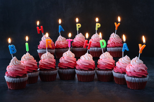 Stock photo showing close-up view of illuminated, brightly coloured individual, letter candles spelling 'Happy Birthday' stuck into rows of freshly baked, homemade, red velvet cupcakes in paper cake cases. The cup cakes have been decorated with swirls of ombre effect pink piped icing.