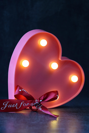 Stock photo showing close-up view of pink, heart shaped box lined with mounted illuminated, light bulbs on a black background. Valentine's Day and romance concept.