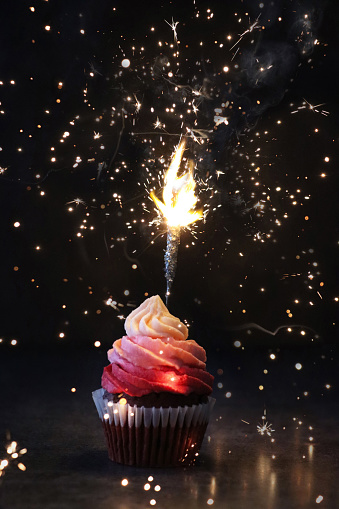 Stock photo showing close-up view of a single freshly baked, homemade, red velvet cupcake in paper cake case with lit, sparkler stuck. The cup cake has been decorated with a swirl of ombre effect pink piped icing.  Valentine's Day and romance concept.