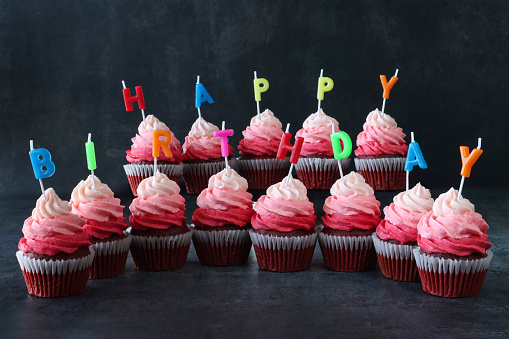 Stock photo showing close-up view of brightly coloured individual, letter candles spelling 'Happy Birthday' stuck into rows of freshly baked, homemade, red velvet cupcakes in paper cake cases. The cup cakes have been decorated with swirls of ombre effect pink piped icing.