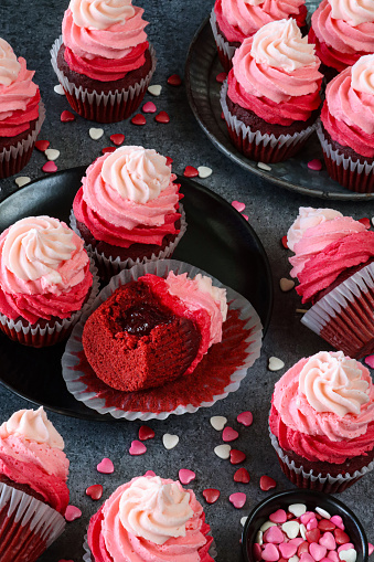 Stock photo showing close-up, elevated view of a batch of freshly baked, homemade, red velvet cupcakes in paper cake cases on and surrounding a plate and cake stand cupcake with unwrapped cake with missing bite showing jam filling. The cup cakes have been decorated with swirls of ombre effect pink piped icing and sugar hearts.  Valentine's Day and romance concept.