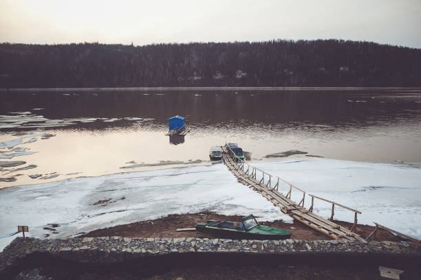 A wooden dock extends into a partially frozen river, with a small boat covered with a blue tarp moored nearby. The serene winter scene is framed by bare trees lining the riverbank.