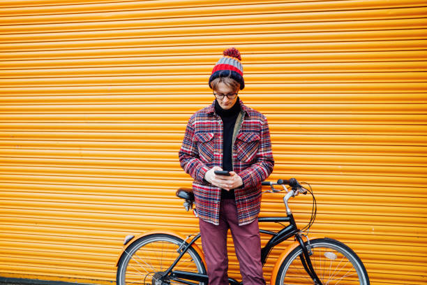 istock Young hipster man using mobile phone while sitting on cruiser bicycle near orange wall. Student using 5g network,rests after riding. Eco friendly transport for carbon footprint. Sustainable lifestyle 1995730606