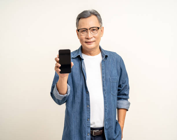istock Handsome asian mature old man using smartphone showing screen display on isolated white background. Portrait of cheerful smiling senior asian man holding cell phone. Mature People and lifestyle 1995808095