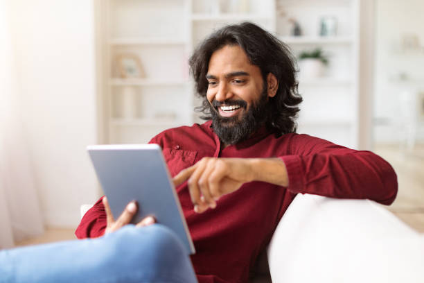 istock Cheerful young indian man sitting on couch, using digital tablet 1995819683