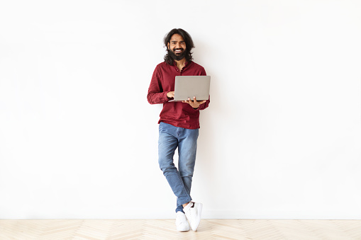 Positive handsome bearded millennial indian guy freelancer independent contractor holding computer laptop, posing on white background, copy space. Online education, remote work