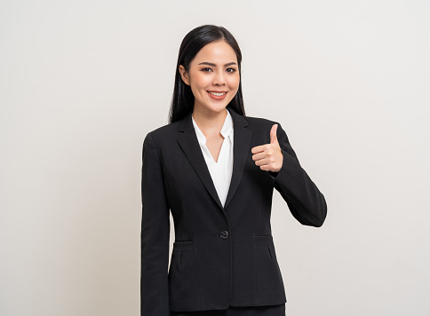 Attractive Young asian business woman thumbs up looking to camera standing pose on isolated white background. Latin Female around 25 in black suit portrait shot in studio.