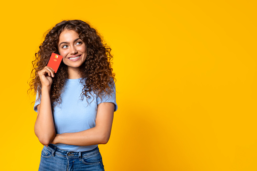 Curly-haired woman holding a credit card to her temple, looking pensive and happy, considering her purchase options on yellow background, free space