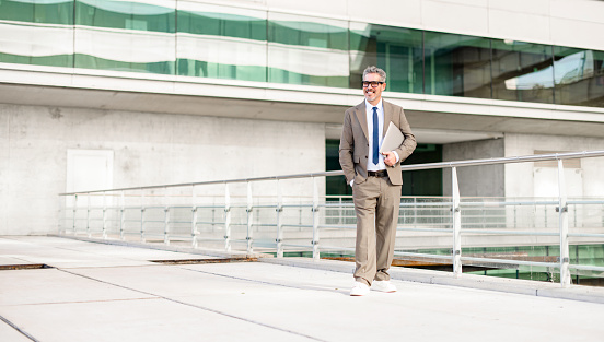 A full-length portrait of a mature grey-haired businessman smiling while walking confidently, laptop in hand, with a modern building in the background, suggesting a dynamic business lifestyle