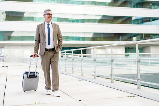 Mature grey-haired businessman strides confidently, suitcase in tow, against an urban backdrop, representing a professional seamlessly blending travel and business