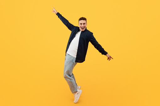 Cheerful young stylish caucasian guy wearing casual clothing moving, standing on toes and stretching hands, posing isolated on yellow background, copy space. Zoomers, gen z lifestyle