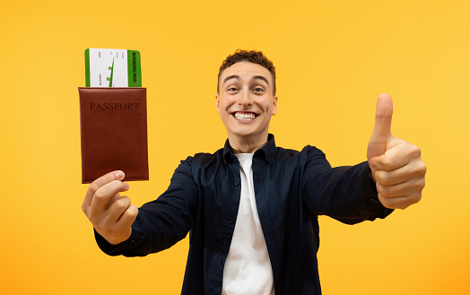 Cheap Tickets Concept. Portrait Of Excited Guy Holding Showing Boarding Pass Passport And Thumb Up, Standing On Yellow Studio Background. Joyful Man Tourist Ready For Travel. Book Your Trip