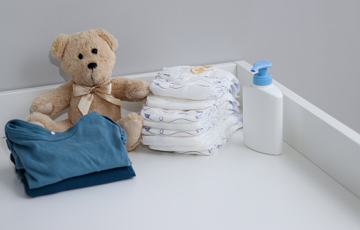 A stack of diapers, teddy bear and baby supplies on white changing table, nursing room