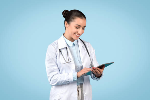 デジタルタブレットを保持し、青い背景の上にオンラインで患者に相談する医療作業服を着たフレンドリーな女性医師 - portrait doctor paramedic professional occupation ストックフォトと画像