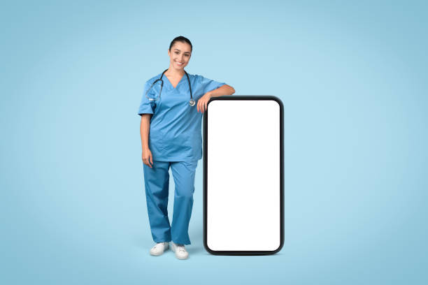 istock Smiling female nurse in blue scrubs leaning on large blank mobile screen 1995923697