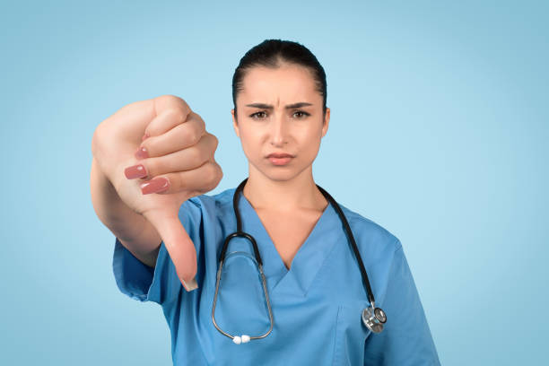 infermiera femminile triste con il pollice in giù su sfondo blu - downbeat foto e immagini stock