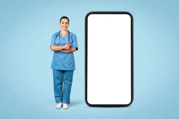 Competent and assured female nurse in blue scrubs with stethoscope, standing arms crossed next to large smartphone with blank screen for copy space