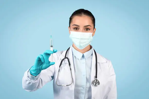 Serious female healthcare professional in a surgical mask and gloves carefully displays a syringe, ready for vaccination or treatment, blue background