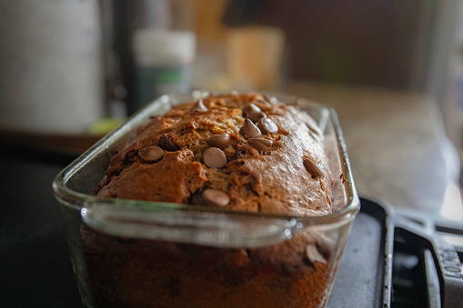 Freshly baked banana bread with chocolate chips sitting on a counter cooling off as the sun shines behind it through a window.