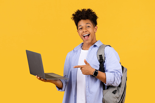 Exuberant african american student with laptop pointing at screen with laugh, possibly sharing digital discovery or success, against bright yellow background