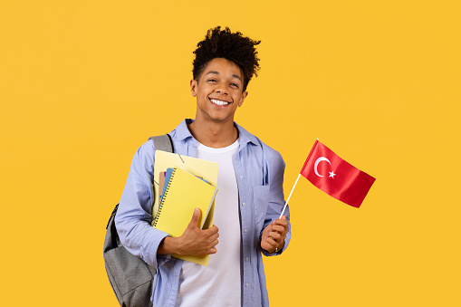 Friendly black scholar with curly hair holding Turkish flag and notebooks, smiling, perhaps for a cultural exchange program, on yellow background