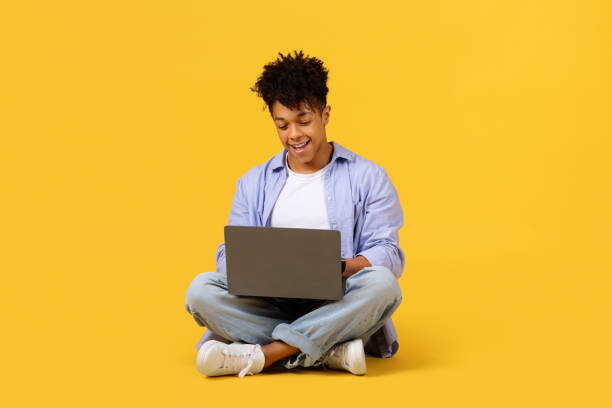istock Engaged black teen guy using laptop on yellow floor 1995947857