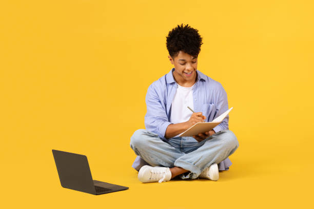 Cheerful black male student writing in notebook beside laptop