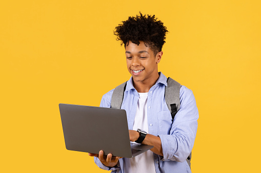 Focused black male student with curly hair using laptop, likely involved in online education or completing assignments, all with pleasant demeanor on yellow background