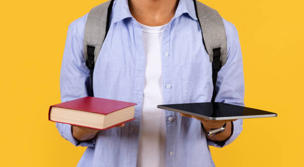 Student male comparing book with tablet on yellow background