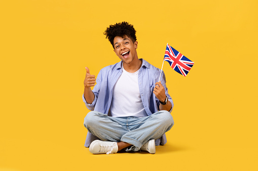 Delighted male student sitting cross-legged, giving thumbs up while holding UK flag, showcasing joyful expression against bright yellow background, signifying success and national pride