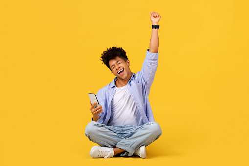 Excited black male student sitting cross-legged, arm raised in triumph, holding smartphone with joyous expression against vivid yellow background, embodying success and positivity