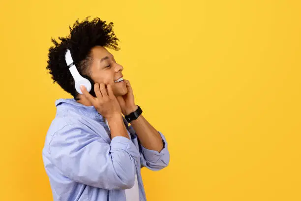 Ecstatic black man with eyes closed, feeling the rhythm while listening to music on headphones, dressed casually with joyful expression, against yellow backdrop, side view, free space