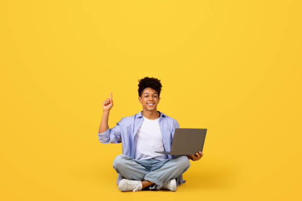 istock Happy black man with laptop points up at free space on yellow 1995948484