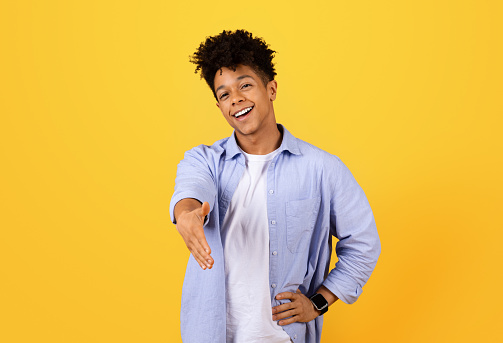 Cheerful young black man with curly hair in casual blue shirt extends friendly handshake against vivid yellow background, embodying warmth and welcome