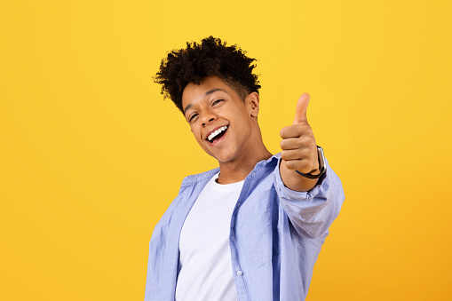 Excited young black male with curly hair flashes thumbs up sign, wearing casual shirt and smartwatch, exuding positivity against vibrant yellow backdrop