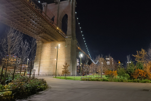 View of underneath the Brooklyn Bridge at night. Photo taken from sidewalks at Brooklyn Bridge Park with bridge lighting shining above.