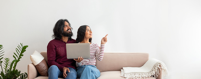Happy millennial indian couple with modern pc laptop in their hands sitting on couch at home in living room, looking at blank space, web-banner for offer, advertisement. Online shopping, banking