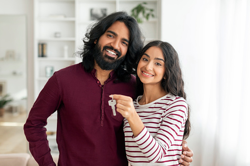 Portrait of happy young indian couple beautiful woman and man hugging and showing key from house, their new apartment, smiling at camera. Millennial family moving to own home. Mortgage, relocation