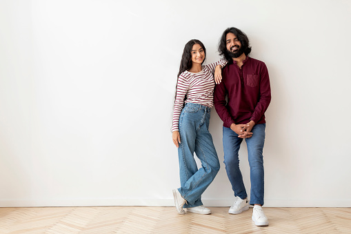 Cheerful beautiful young indian couple stylish man and woman wearing casual outfit posing on white blank wall background, embracing and smiling at camera, copy space. Moving, relocation