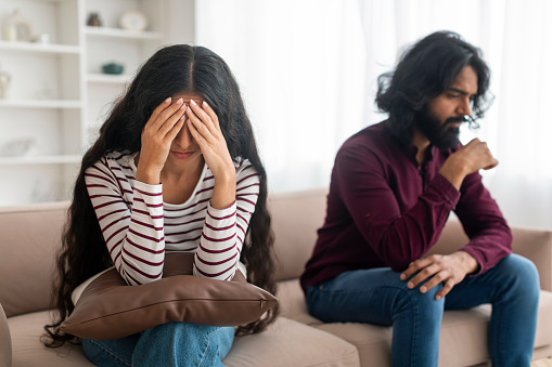 Frustrated young indian couple sitting on couch after quarrel at home. Unhappy millennial spouses experiencing difficulties crisis in marriage, have fight, living room interior