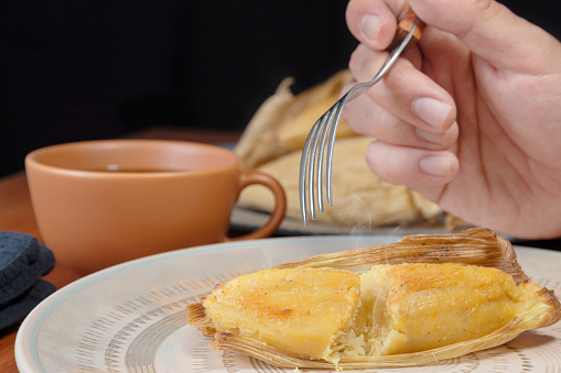 Hand of an unrecognizable person eating a hot ¨Envuelto de Mazorca¨ accompanied by a cup of coffee. Traditional Colombian corn meal. Wrapped corn. Breakfast in Colombia.
